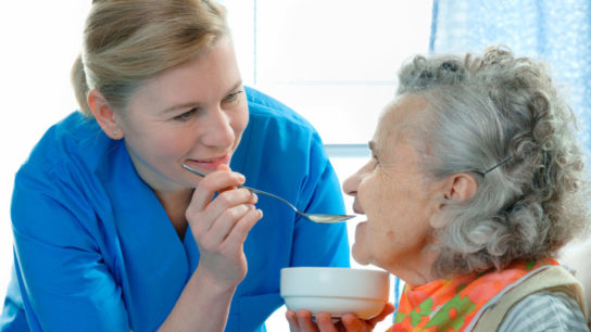 Image of an elderly nursing home resident receiving help with feeding by a nursing assistant