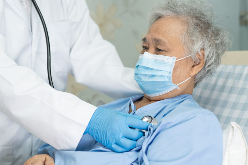 Sick elderly person wearing surgical face mask and being cared for by a clinician with stethoscope