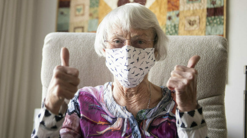 Senior woman wearing surgical face mask sits in chair while giving the thumbs-up sign