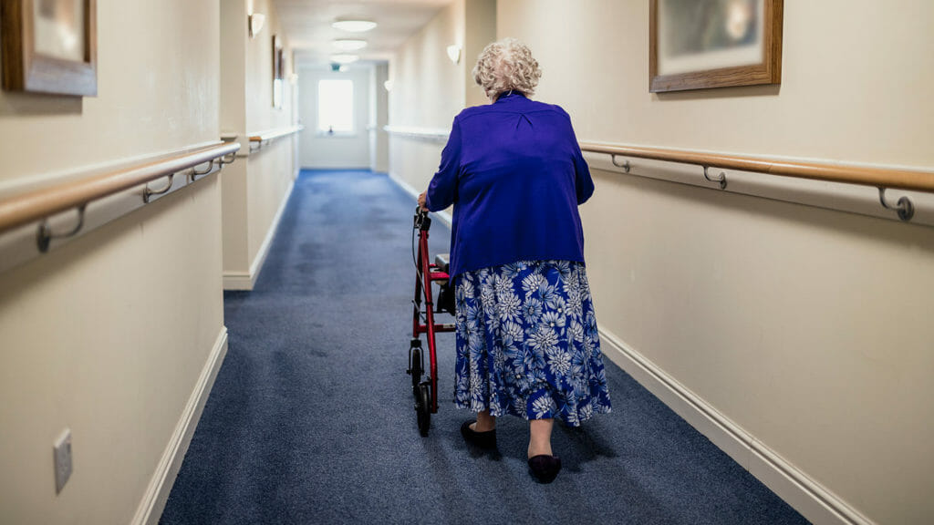 A nursing home resident walks down an empty hallway with her walker