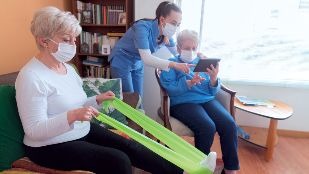 A nurse helps a nursing home resident with her tablet while another resident to the left of her stretches her legs