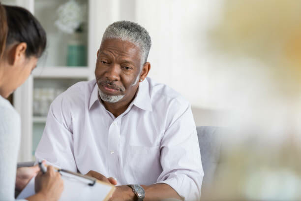 A senior man sits on a couch opposite his unrecognizable female therapist and looks down with a sad expression.  His therapist takes notes with a pen and clipboard.