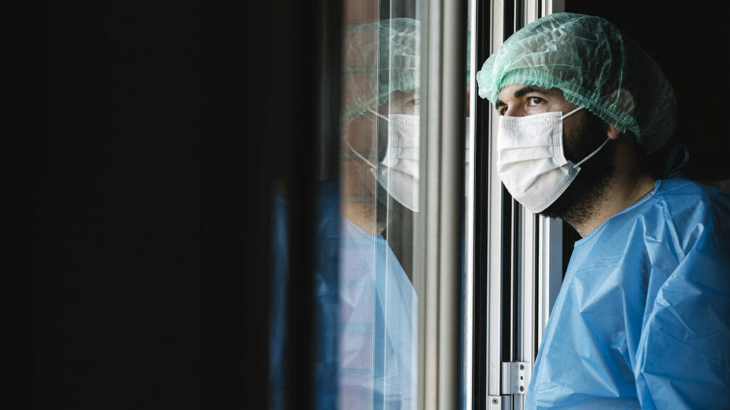 Doctor in face mask is looking away through window with tiredness while having a break