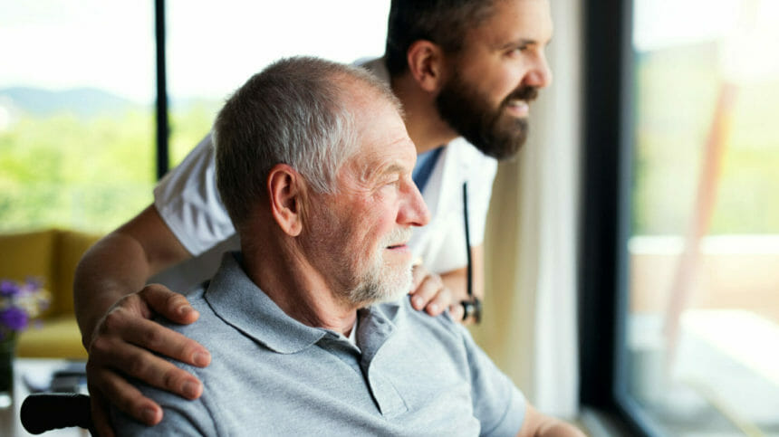 A mature man caregiver with stethoscope and older, senior patient looking out through window.