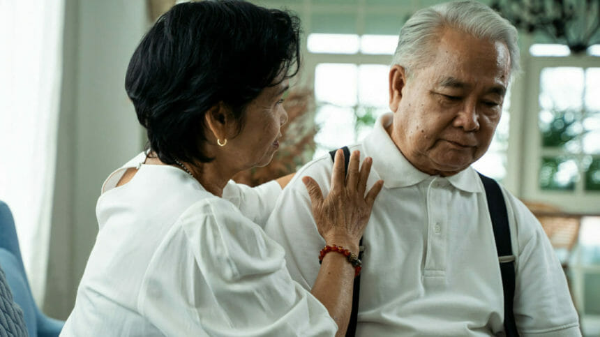Asian senior couple sitting on the sofa while elderly wife comforts her sad husband at home.
