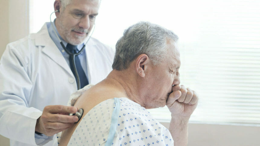 Male doctor using stethoscope to examine coughing patient in hospital gown.