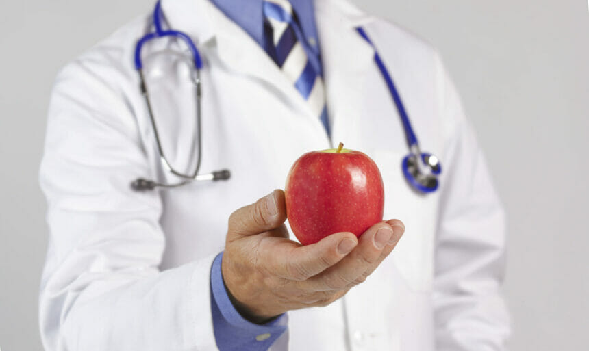 Image of a doctor in white coat and stethoscope holding an apple to camera