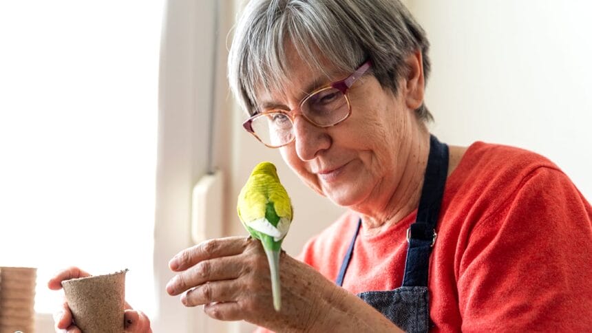 Older woman plants vegetable seeds while she holds a parakeet.